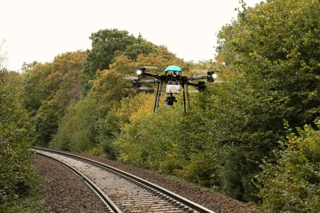 A drone equipped with a camera flying over railway tracks surrounded by green foliage.