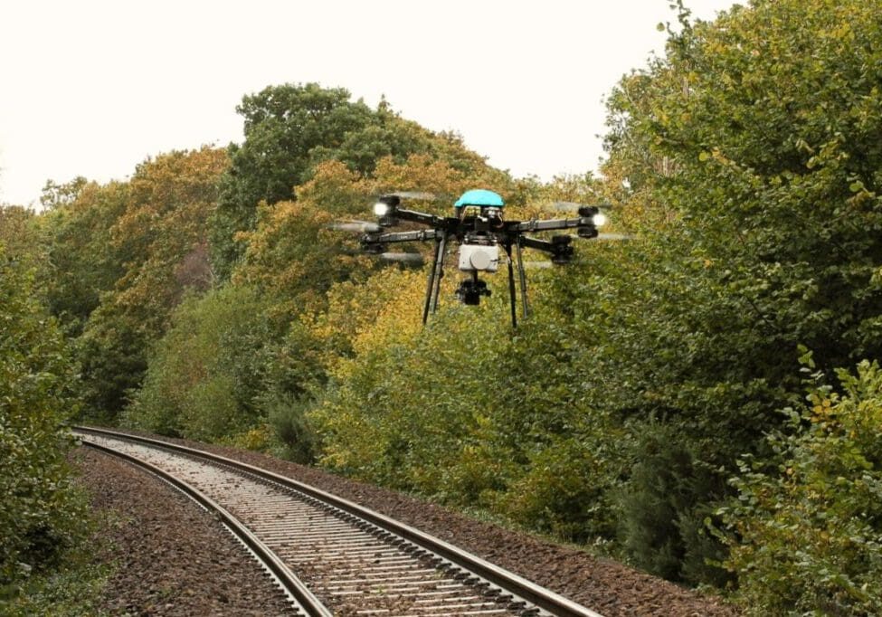 A drone equipped with a camera flying over railway tracks surrounded by green foliage.